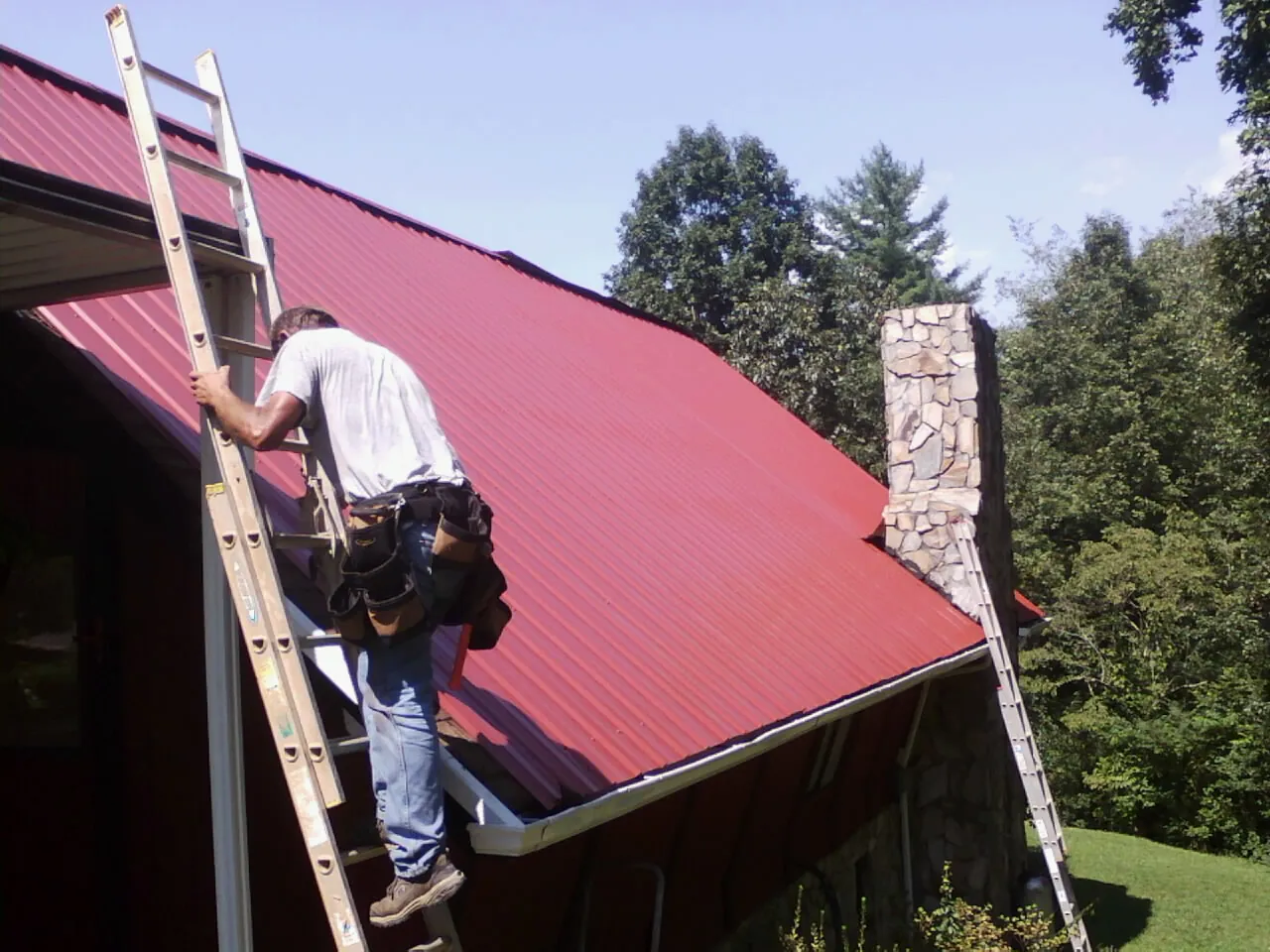 Roofer on a ladder beside a red metal roof and stone chimney — Soffit & Fascia Repair by Roofing Brothers in Wauseon